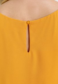 Gele blouse achterkant met een keyhole-opening en een ronde knoopsluiting bij de hals, gemaakt van een zachte stof met een gladde textuur.
