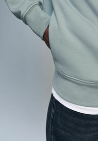 Felpa verde chiaro con finitura testurizzata, polsini aderenti e tasca laterale. Abbinata a uno strato bianco sottostante e jeans scuri.