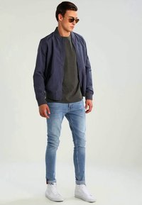 Chaqueta bomber navy, camiseta gris, jeans azul claro y zapatillas blancas. Gafas de sol reflectantes. Look casual con un diseño ajustado.