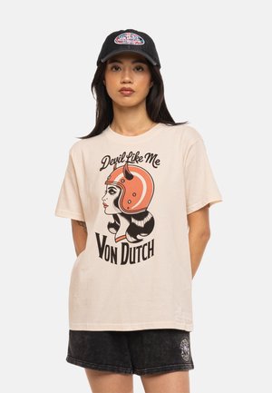 Femme portant un t-shirt beige Von Dutch avec une silhouette féminine en casque orange et short noir, portant également une casquette noire Von Dutch.