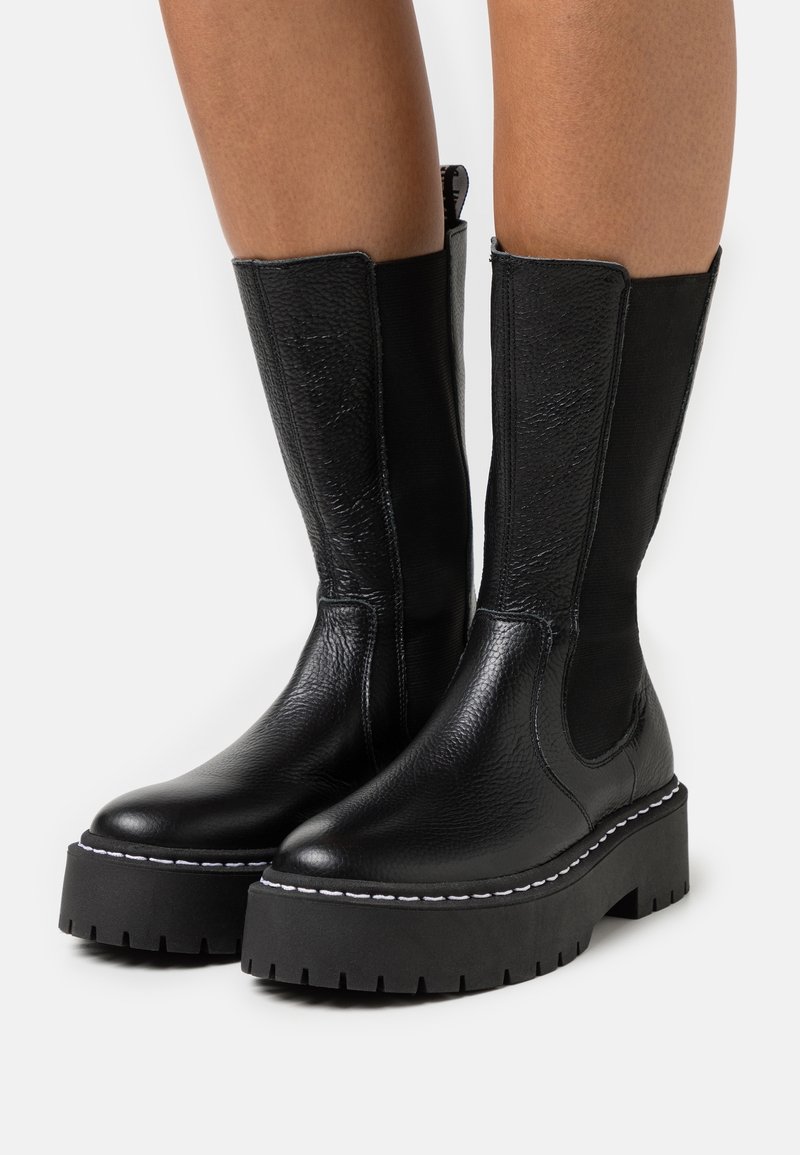 Bottes en cuir noires mi-mollet avec semelles épaisses à plateforme et coutures blanches portées sur des jambes nues sur un fond blanc.