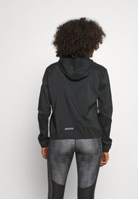 Musta kevyt takki hupulla, jossa on joustavat hihat ja helma. Yhdistettyä kuvioitujen legginsien kanssa, joissa on toistuva logodesign.