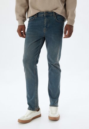 Jeans de denim azul de pierna recta con un acabado ligeramente desvaído, cinco bolsillos y cierre de botón. Llevados con zapatillas blancas y marrón claro.