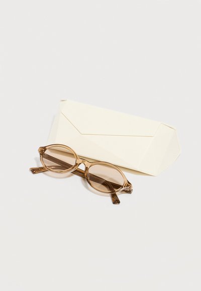 Στρογγυλά καφέ διαφανή γυαλιά ηλίου τοποθετημένα μπροστά από μια κλειστή μπεζ ορθογώνια θήκη γυαλιών πάνω σε λευκή επιφάνεια.