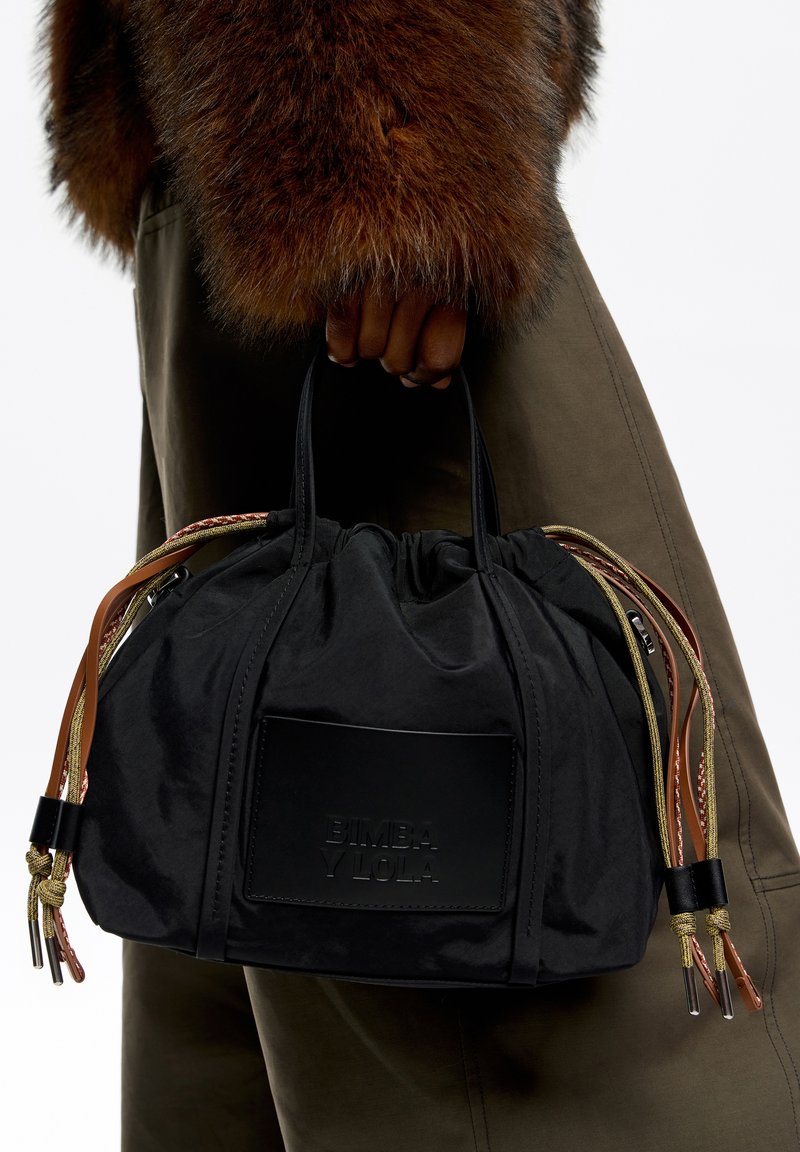 Bolsa de tela negra con parte superior recogida, con asas trenzadas en marrón y dorado, logo en relieve y superficie texturizada.