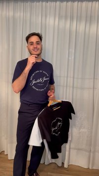 Muž drží dve tričká na vešiakoch, má na sebe tmavomodrú košeľu Jack & Jones, stojí pred bielymi závesmi v miestnosti.