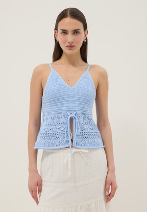Frau trägt ein hellblaues gehäkeltes ärmelloses Oberteil mit Bändern an der Taille und einen weißen Rock mit Spitzenbesatz, stehend vor einfachem Hintergrund.