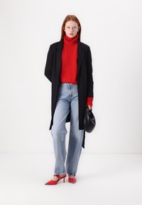 Long manteau noir, col roulé rouge, jeans bleu clair, escarpins rouges avec accents argentés et un sac à main noir. Fond simple.