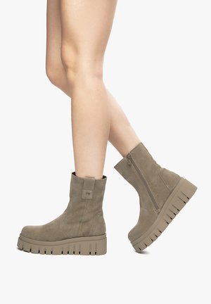 Brown suede ankle boots with a thick, ridged sole and a side zipper. The design features a pull tab at the back for easy wear.