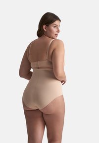 Donna curvy che indossa biancheria modellante beige a vita alta e reggiseno abbinato, in piedi con la schiena e la testa girate di lato.