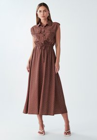 Brown, sleeveless maxi dress with a button-up front and drawstring waist. White polka dot pattern throughout. Smooth fabric, casual design.