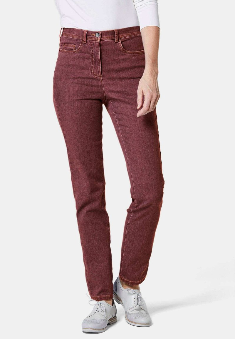 Jean skinny en denim rouge avec cinq poches, fermeture à bouton à l'avant et coutures contrastées ; porté avec des chaussures de ville gris clair.