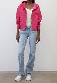 Persona con una sudadera con cremallera de color rosa brillante, camiseta sin mangas blanca, jeans de campana azul claro y zapatillas blancas de pie contra una pared blanca.