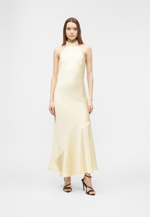 Naine, kes kannab varrukateta kreemjat satiinsest poolkaeluse ja asümmeetrilise äärega kleiti ning mustade rihmikuga kõrgete kontsadega sandaale, seisab ühevärvilise tausta ees.