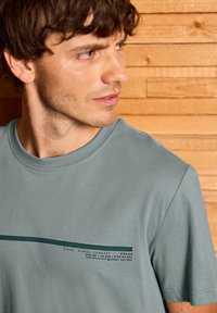 T-shirt en coton gris clair avec un col rond, des manches courtes et un imprimé graphique vert avec un texte sur la poitrine.