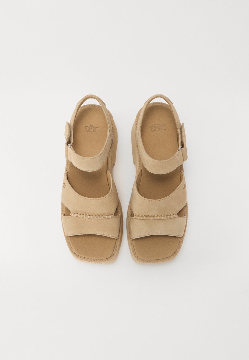 Beige Wildledersandalen mit flacher Sohle, die zwei breite Riemen über dem Fuß und einen verstellbaren Knöchelriemen für einen sicheren Sitz besitzen.