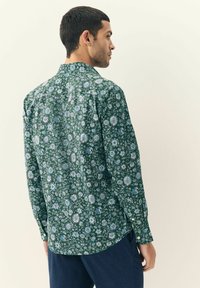 Mand iført en grøn langærmet skjorte med blomster i blå og hvide nuancer, vender sidelæns, i kombination med mørkeblå bukser.