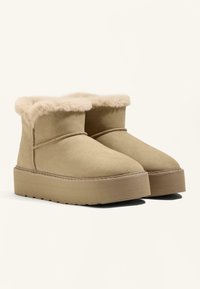 Bež nubuk ankle čizme s plišanim krznom na rubu, ravnim platformama, zaobljenim vrhom i suptilnim šavnim detaljem duž gornjeg ruba.