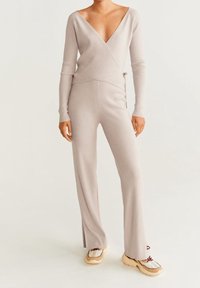 Beiger, gerippter Jumpsuit mit tiefem V-Ausschnitt, langen Ärmeln und ausgestellten Beinen. Auffällige seitliche Schlitze, kombiniert mit weißen und hellbraunen Sneakers.