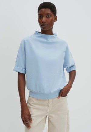 Donna che indossa una blusa a maniche corte di colore azzurro chiaro con collo alto e pantaloni beige, in piedi con una mano in tasca su uno sfondo semplice.