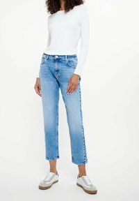 Person som bär ljusblå raka jeans, vit långärmad tröja och vita sneakers med grå detaljer mot en neutral bakgrund.