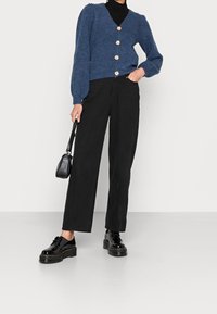 Cardigan bleu tricoté avec un col en V et cinq gros boutons, assorti à un pantalon noir à jambes larges et des chaussures noires vernies à lacets. Sac à main noir.
