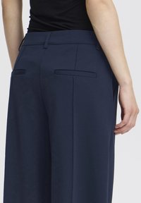 Vedere din spate a unei persoane purtând pantaloni bleumarin închis cu bucle pentru curea și buzunare haklate la spate, asortați cu un top negru mulat.