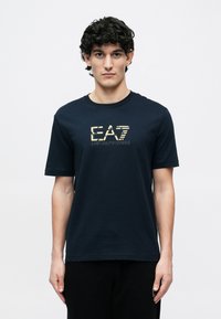 Sötétkék pamut póló aranyszínű, elöl nyomtatott "EA7 EMPORIO ARMANI" logóval, rövid ujjakkal és kerek nyakkivágással.