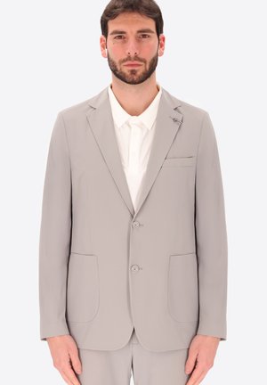 Giovane uomo con capelli corti scuri e barba, indossa una giacca da abito grigio chiaro sopra una camicia bianca con colletto, in piedi davanti a uno sfondo chiaro e uniforme.