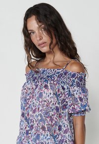 Blusa floreale off-shoulder con scollatura arricciata, maniche corte e un leggero tessuto trasparente con motivi rosa e blu.