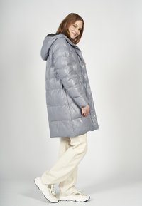 Grijze pufferjas met een glanzende afwerking, capuchon en horizontaal doorgestikt ontwerp. Witte broek en lichte sneakers maken de outfit compleet.