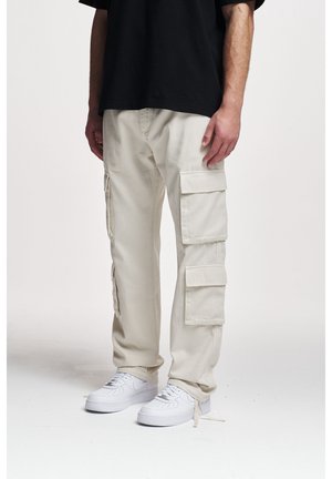 Pantalones cargo beige con múltiples bolsillos laterales, ajustados en los tobillos, combinados con zapatillas blancas y una camiseta negra de gran tamaño.