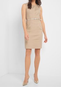 Robe beige sans manches avec une silhouette slim et une fine ceinture blanche. Texture lisse, longueur genou, design minimaliste, portée avec des talons métalliques.