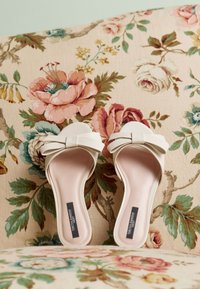 Mules ouvertes crème avec un détail nœud, présentant une texture douce et un design léger, posées sur un fond en tissu à motifs floraux.