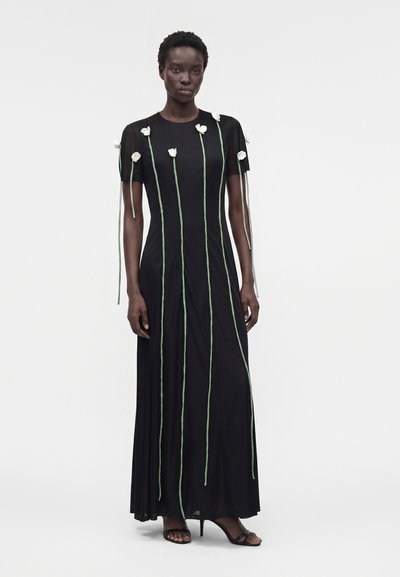 Γυναίκα που φοράει μακριά μαύρη φορεσιά με κοντά μανίκια, διακοσμημένη με λευκές εφαρμογές λουλουδιών και πράσινους μίσχους που εκτείνονται κάθετα.