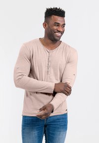 Langärmliges, hellbeiges Henley-Shirt aus weichem Stoff, mit Knopfleiste und detaillierten Nähten an den Ärmeln. Kombiniert mit blauen Jeans.