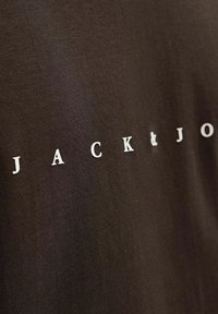 Brun bomulds T-shirt med teksten "JACK & JONES" i hvide bogstaver, placeret centralt med en glat, mat tekstur.