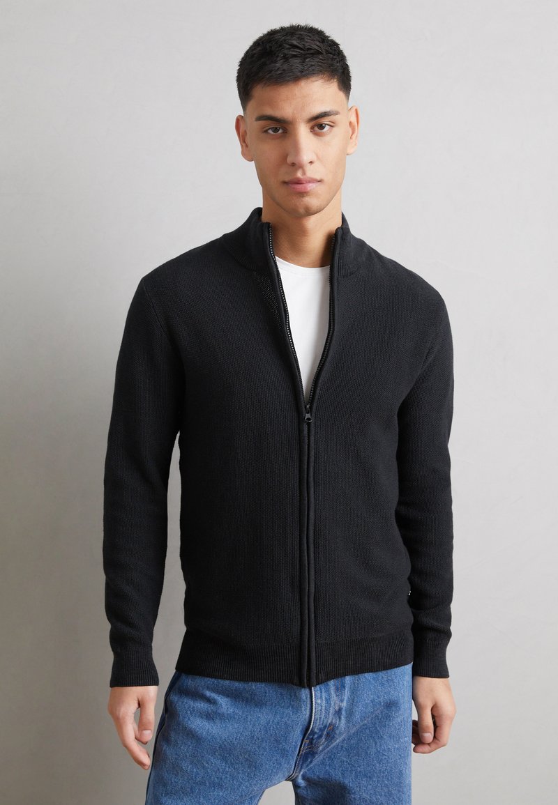 Svart glidelås-cardigan i teksturert strikk, med høy krage og ribbestrikkede mansjetter, båret over en hvit t-skjorte og løse blå jeans.