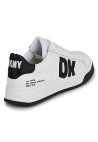 Zapatilla de cuero blanca con acento negro en el talón y letras "DK" en negrita. Presenta una superficie texturizada y una suela de goma negra. Texto en el detalle del talón.