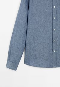 Chemise chambray bleu à manches longues avec des boutons blancs et une seule manchette sur un fond blanc uni.