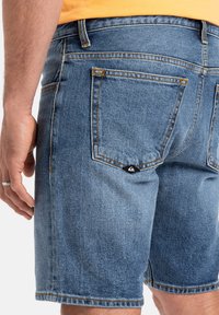 Pantalones cortos de mezclilla en azul claro con costuras visibles y un acento de diseño en el bolsillo trasero, que destacan la textura y las características de diseño casual.