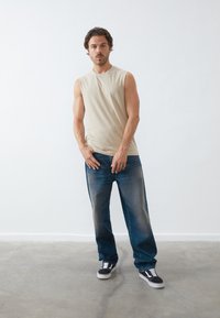 Beige sleeveless t-shirt, loose blue jeans with light fading, and black sneakers with white accents. Model stands against a plain background.