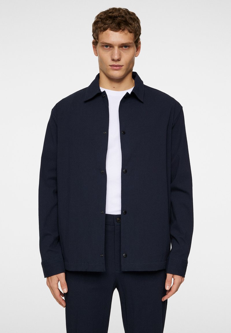 Marineblauwe button-up jas, gemaakt van gestructureerde stof met een kraag en voorzakken, gedragen over een wit T-shirt.