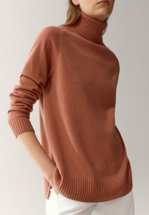 Femme portant un pull à col roulé couleur rouille avec poignets et ourlet côtelés, associé à un pantalon blanc, main dans la poche.