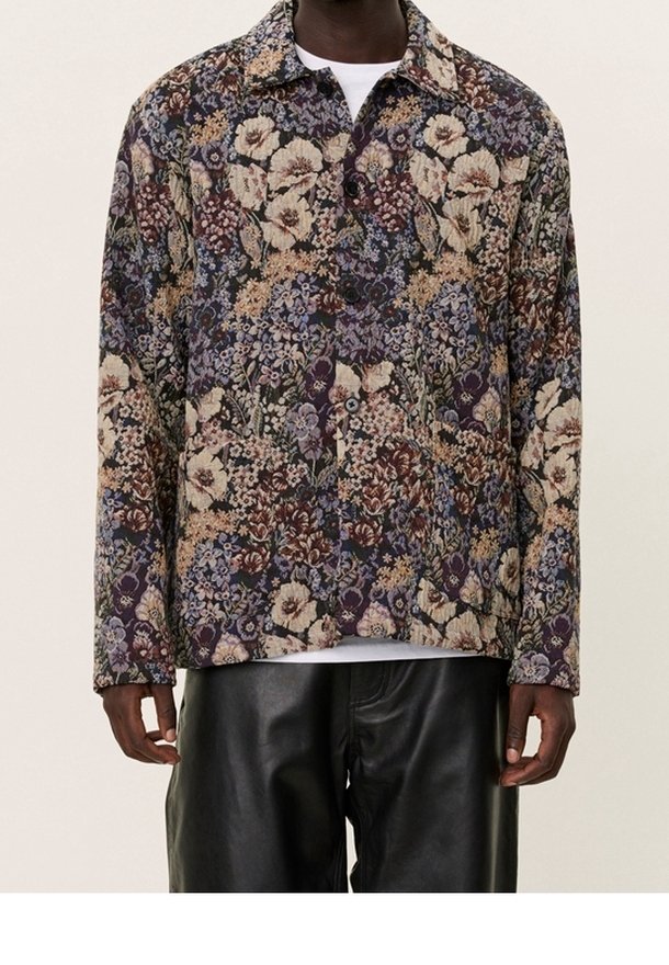 Homme portant une veste boutonnée à motif floral dans des couleurs sourdes sur une chemise blanche, associée à un pantalon en cuir noir.
