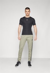 Sort tætsiddende atletisk skjorte, olivengrønne bukser med sorte side-striber, båret med sort og hvidt sneakers; glat tekstur, minimalistisk design.