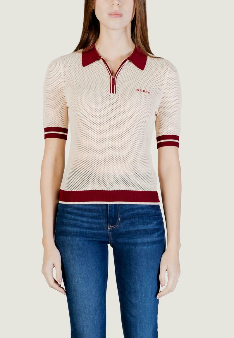 Femme portant un polo en maille beige avec un col, des poignets de manches et un ourlet rouges, associée à un jean bleu, debout devant un fond uni.