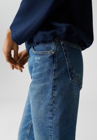 Persona che indossa un maglione blu a trama e jeans blu sbiaditi con vita regolabile, con le mani rilassate lungo i fianchi.