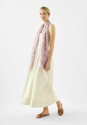 Vrouw gekleed in een mouwloze crèmekleurige maxi-jurk, bruine sandalen met open tenen en een lange roze sjaal met paisleyprint, staand tegen een witte achtergrond.