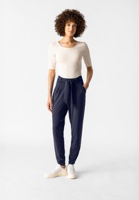 Beige figurnära topp med korta ärmar, kombinerad med högmidjade marinblå byxor med en knytdetalj. Modellen bär vita sneakers.